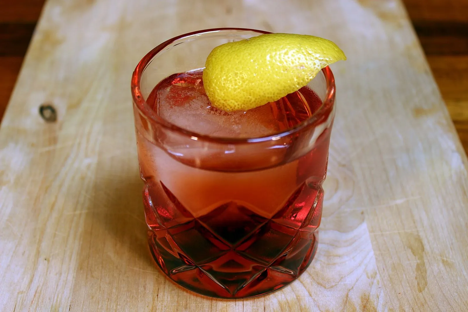 Cocktail rosso in un bicchiere di cristallo con ghiaccio e scorza di limone su un tagliere di legno